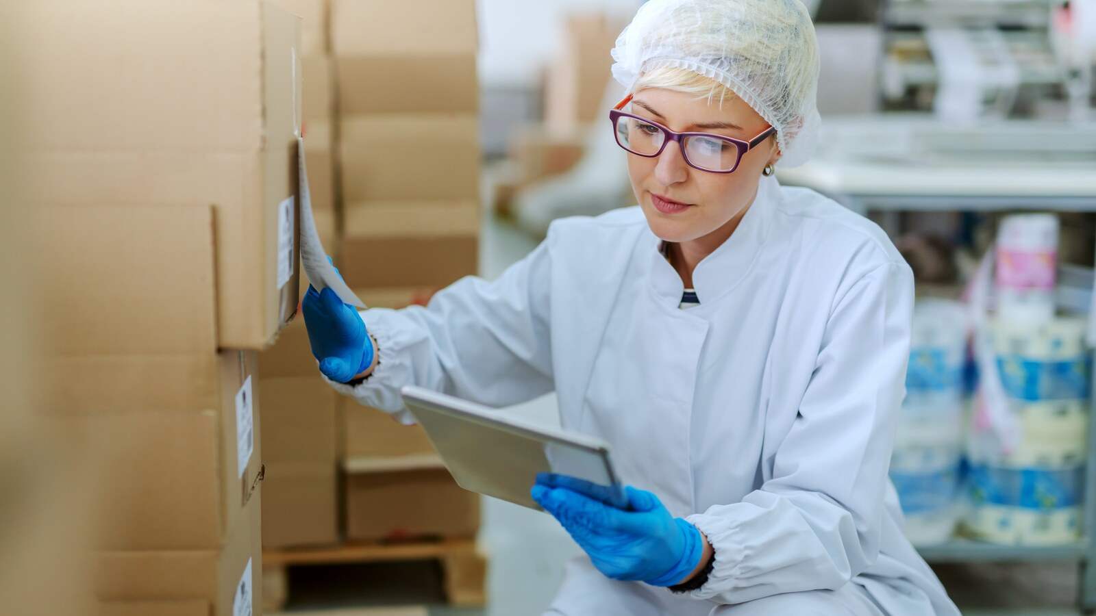 Técnica de una planta alimentaria con cofia y guantes revisando con una tableta las etiquetas de lote de las cajas de producto
