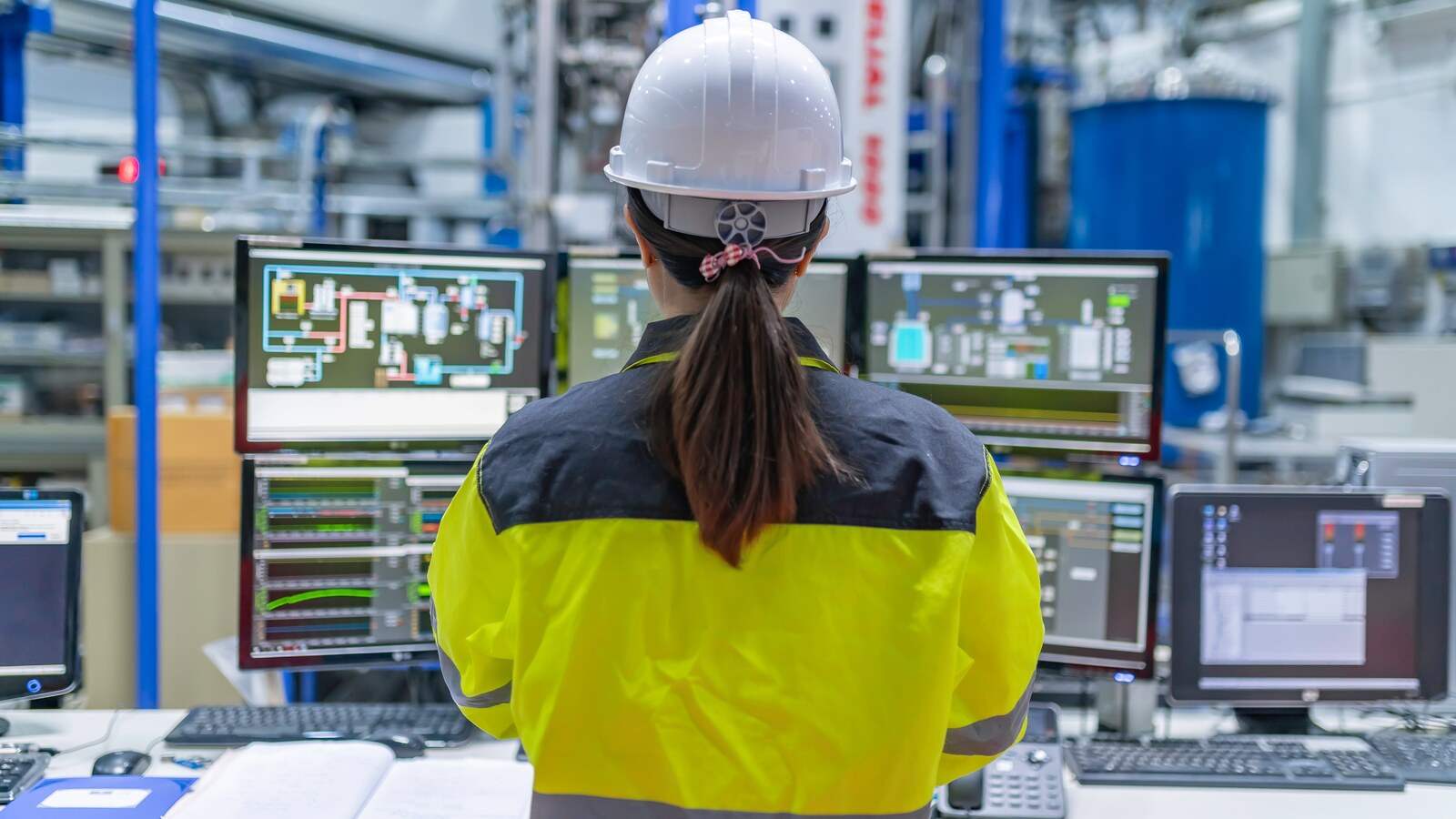 Técnica con casco y chaleco de alta visibilidad frente a las pantallas SCADA de un puesto de control dentro de la planta