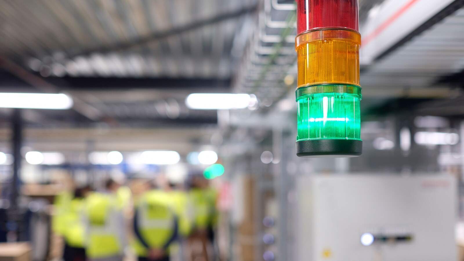 Torre de señalización luminosa con la luz verde encendida sobre una línea de producción, con operarios con chaleco al fondo